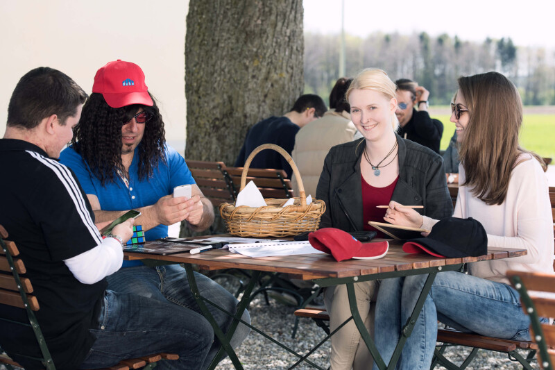 Vier Personen am Tisch draußen. Baumstamm im Hintergrund